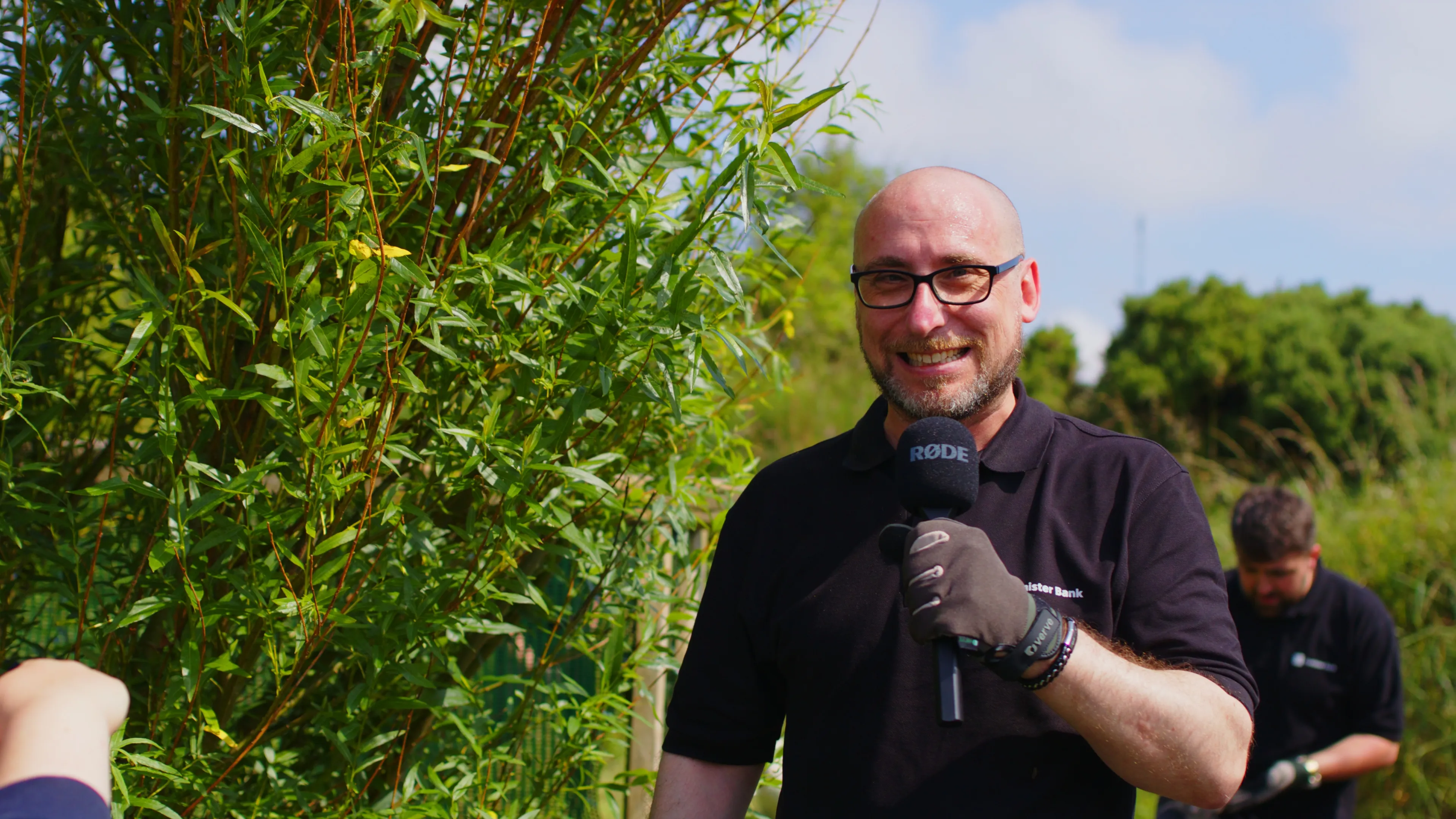 A man in glasses and a black shirt smiles while holding a microphone in a lush garden. Sunlit greenery surrounds him, creating a vibrant, cheerful atmosphere.